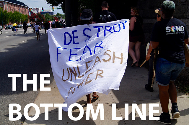 The words "The Bottom Line" appear over a group of people walking and carrying a sign that reads "Destroy Fear, Unleash Power"